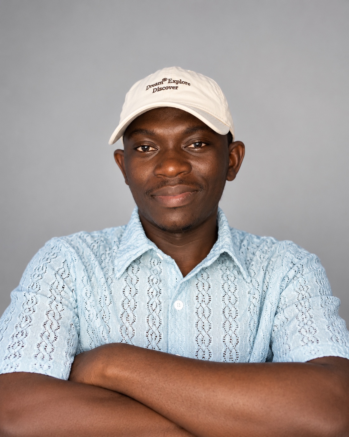 Mojeeb Titilayo, founder of Arcapush, smiling with arms folded. He wears a cream cap and a light-blue patterned short-sleeve shirt against a grey studio backdrop.
