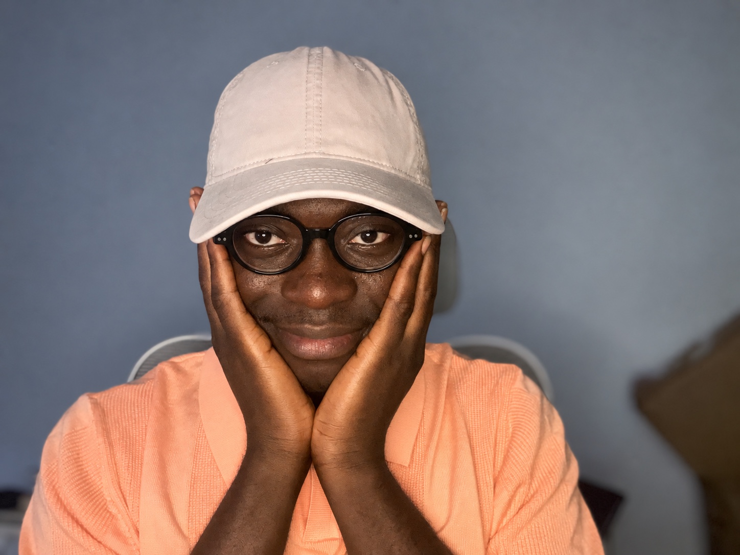 Mojeeb Titilayo smiling at the camera with both hands against his cheeks. He wears a light cap, round black glasses and a peach shirt.